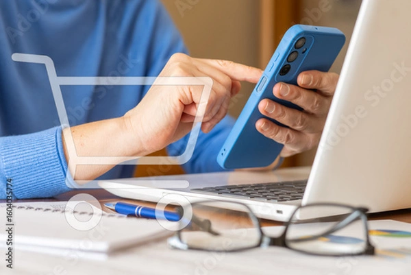 Fototapeta Businesswoman using smartphone for online shopping with a virtual cart icon, while evaluating kpi performance data on laptop with documents, pen and eyeglasses on desk