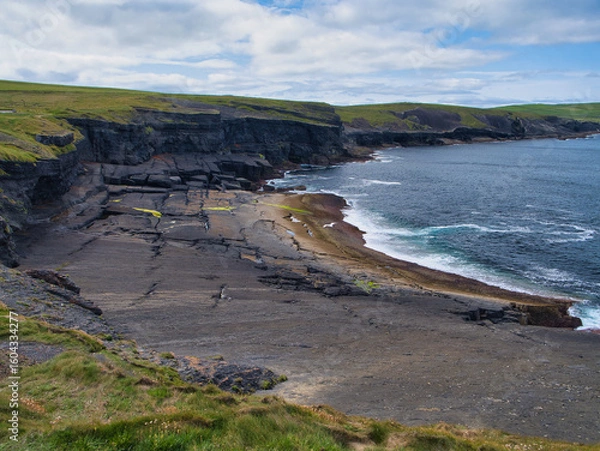 Fototapeta Breathtaking view of Kilkees coastal cliffs, highlighting the striking stratified rock formations against the ocean backdrop under a cloudy sky.