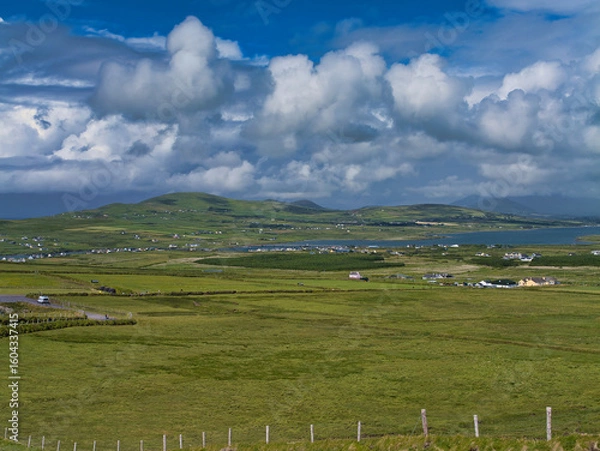 Fototapeta A panoramic view of rolling green fields and scattered cottages under a dramatic cloudy sky in Portmagee, Ireland.