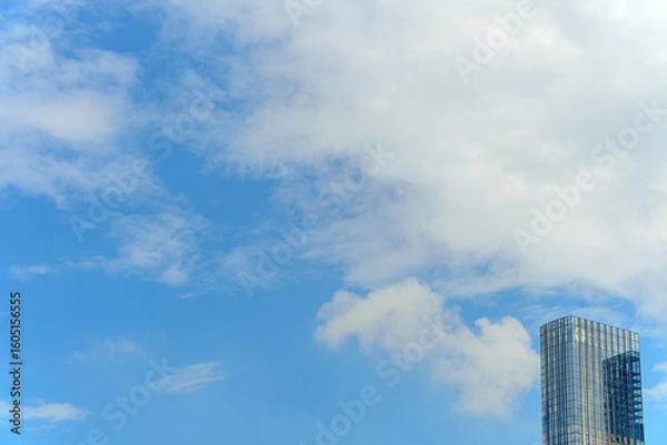 Fototapeta  A modern tall building with mirrored glass reflects the cloudy sky, evoking a sense of harmony between architecture and nature. Its elegant silhouette rises above the urban landscape.