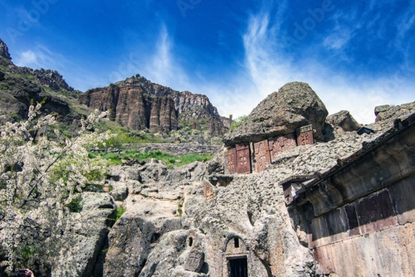Fototapeta Geghard Monastery carved in rock, surrounded by basalt cliffs and blooming trees in spring, Armenia UNESCO heritage site