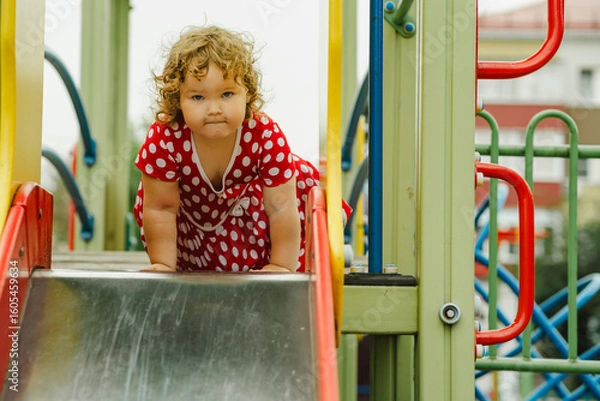 Obraz A young child wearing a red polka dot dress and yellow shoes plays happily on a playground slide The atmosphere is cheerful and colorful showcasing an enjoyable day outdoors