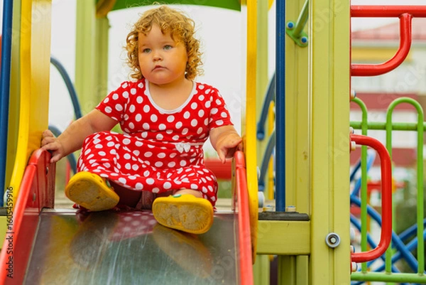 Obraz A young child wearing a red polka dot dress and yellow shoes plays happily on a playground slide The atmosphere is cheerful and colorful showcasing an enjoyable day outdoors
