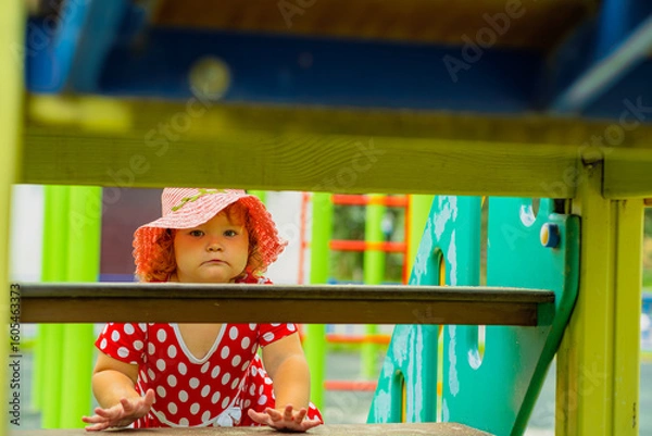 Obraz A young child is climbing through a playground structure on a sunny day Dressed in a red dress with white polka dots and a matching hat she explores with curiosity