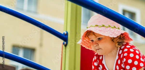 Obraz A young child wearing a red polka dot dress and a hat is climbing on colorful playground equipment The sun is shining in a vibrant outdoor park setting creating a joyful atmosphere
