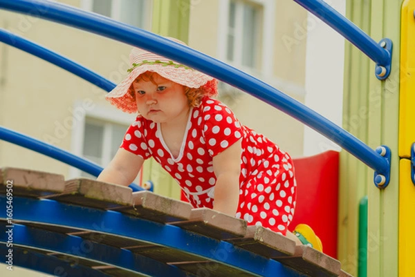Obraz A young child wearing a red polka dot dress and a hat is climbing on colorful playground equipment The sun is shining in a vibrant outdoor park setting creating a joyful atmosphere