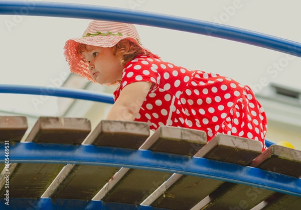 Obraz A young child wearing a red polka dot dress and a hat is climbing on colorful playground equipment The sun is shining in a vibrant outdoor park setting creating a joyful atmosphere