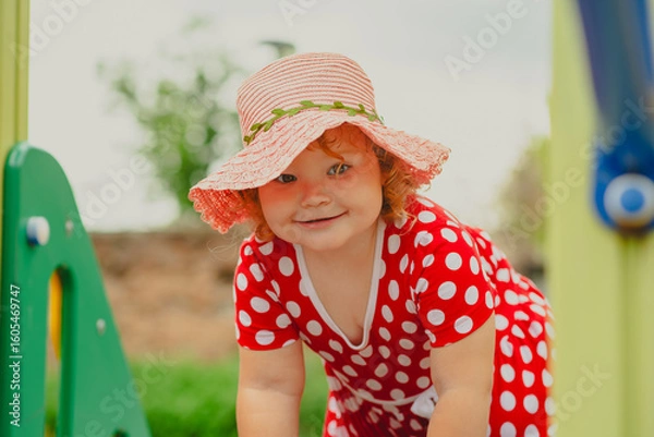 Obraz A young child wearing a red polka dot dress and a hat is climbing on colorful playground equipment The sun is shining in a vibrant outdoor park setting creating a joyful atmosphere