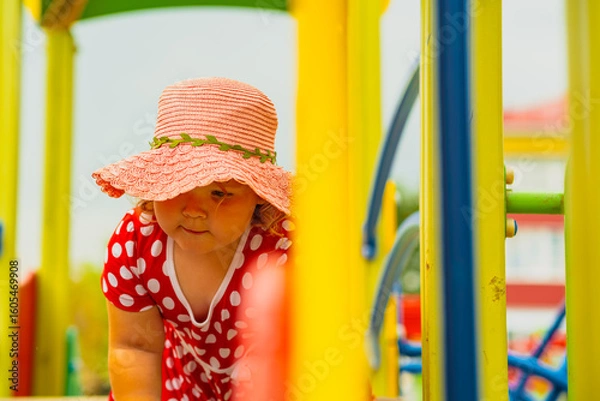 Obraz A young child wearing a red polka dot dress and a hat is climbing on colorful playground equipment The sun is shining in a vibrant outdoor park setting creating a joyful atmosphere