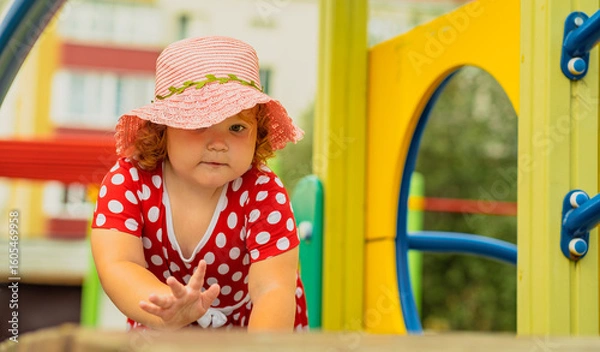 Fototapeta A young child wearing a red polka dot dress and a hat is climbing on colorful playground equipment The sun is shining in a vibrant outdoor park setting creating a joyful atmosphere