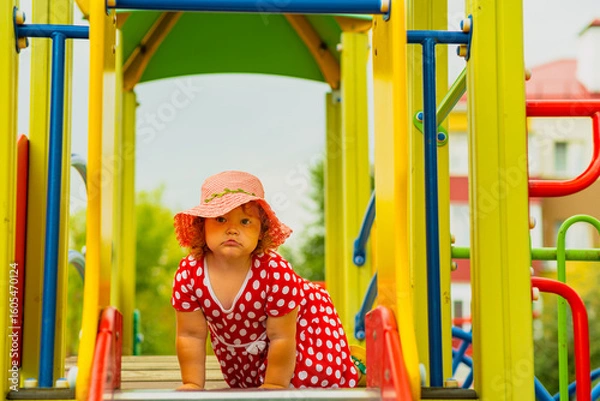 Obraz A young child wearing a red polka dot dress and a hat is climbing on colorful playground equipment The sun is shining in a vibrant outdoor park setting creating a joyful atmosphere