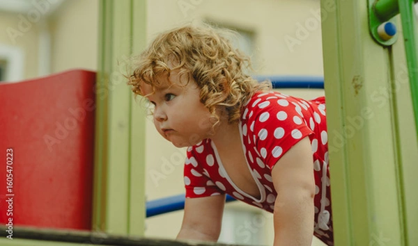 Obraz A young child wearing a red polka dot dress and a hat is climbing on colorful playground equipment The sun is shining in a vibrant outdoor park setting creating a joyful atmosphere