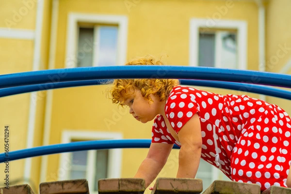 Fototapeta A young child wearing a red polka dot dress and a hat is climbing on colorful playground equipment The sun is shining in a vibrant outdoor park setting creating a joyful atmosphere