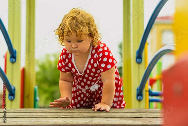 Fototapeta A young child wearing a red polka dot dress and a hat is climbing on colorful playground equipment The sun is shining in a vibrant outdoor park setting creating a joyful atmosphere