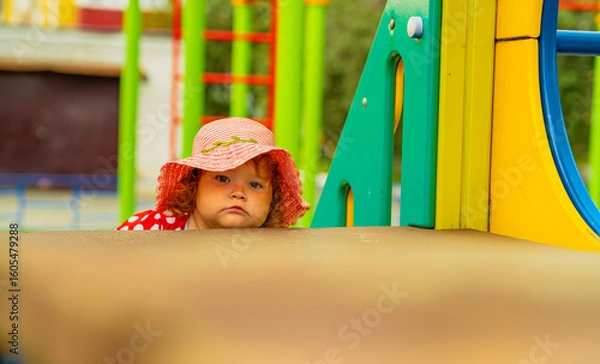 Obraz A young child with curly red hair and a pink hat peers out from behind brightly colored playground equipment It is a sunny day perfect for outdoor fun and exploration