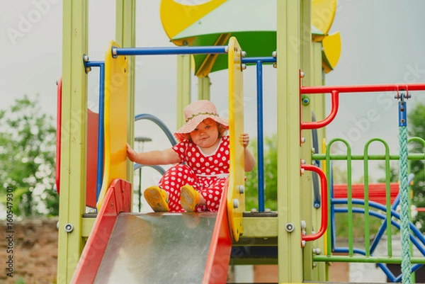 Obraz A happy kid in a red polka dot dress is sliding down a playground slide Bright colors surround her and the atmosphere is joyful during a sunny day with playful clouds
