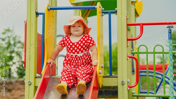 Obraz A happy kid in a red polka dot dress is sliding down a playground slide Bright colors surround her and the atmosphere is joyful during a sunny day with playful clouds