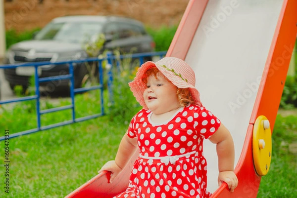 Obraz A happy kid in a red polka dot dress is sliding down a playground slide Bright colors surround her and the atmosphere is joyful during a sunny day with playful clouds