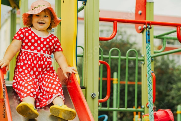 Obraz A happy kid in a red polka dot dress is sliding down a playground slide Bright colors surround her and the atmosphere is joyful during a sunny day with playful clouds