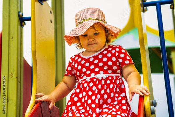 Obraz A happy kid in a red polka dot dress is sliding down a playground slide Bright colors surround her and the atmosphere is joyful during a sunny day with playful clouds
