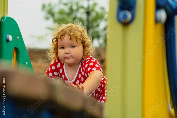 Obraz A young child wearing a red polka dot dress and a hat is climbing on colorful playground equipment The sun is shining in a vibrant outdoor park setting creating a joyful atmosphere
