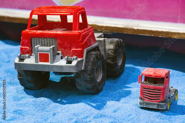 Obraz Two toy trucks are placed in a sandy play area A large red truck stands prominently next to a smaller red truck creating a playful contrast in size and inviting imaginative play for children