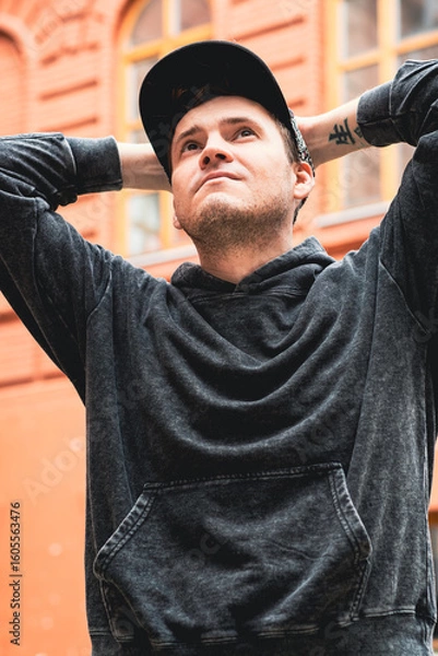Obraz A man stands with hands on his head wearing a black hoodie and cap He looks contemplative in front of a textured red brick wall on a sunny day