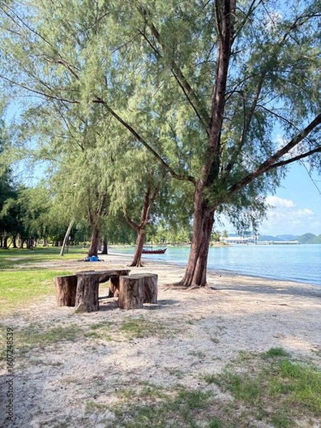 Fototapeta A peaceful beach scene with a wooden table and seats under tall trees, overlooking calm blue waters and distant mountains, ideal for a tranquil escape in Malaysia.