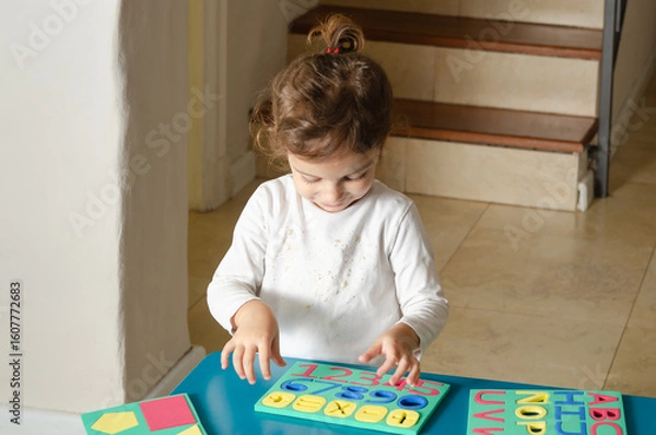Fototapeta A little girl playing with colorful foam letters, numbers and geometric shapes on a blue table. 