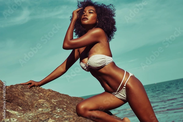 Obraz Egzotyczna modelka z opaloną skórą i kręconymi włosami w bikini pozuje na kamienistej plaży