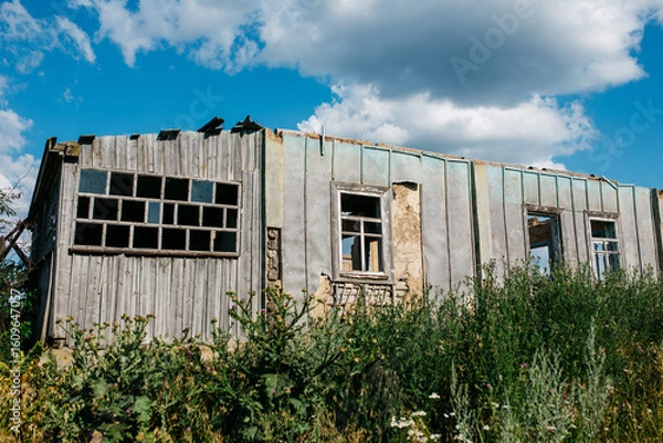 Fototapeta An old wooden abandoned house with broken windows stands in a field among overgrown grass on a summer day against a clear and clear blue sky with clouds. the texture of an old house