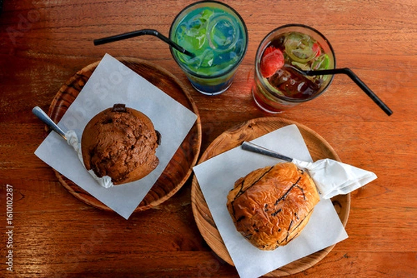 Fototapeta Top view of chocolate muffin, sweet bread, and colorful iced drinks on wooden table. Delicious snack and beverage setup in a cozy café atmosphere.