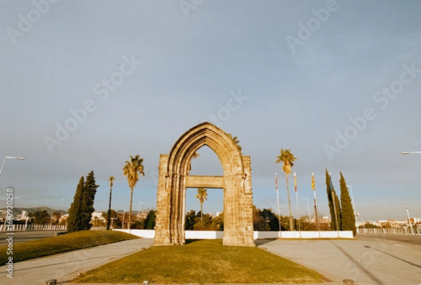 Fototapeta A color analog 35mm film photograph captures an ancient stone arch monument in a Barcelona roundabout, surrounded by palm trees under a bright sky, with classic film grain.