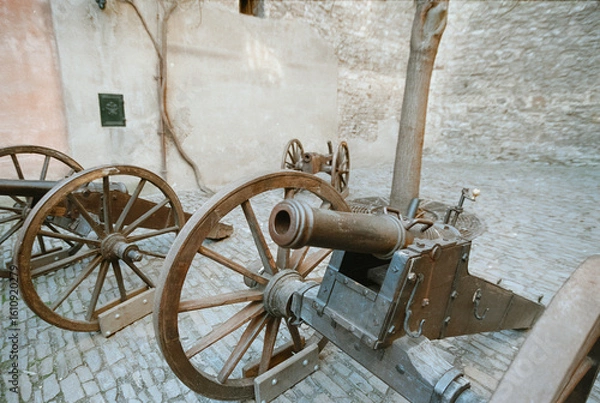 Fototapeta An analog 35mm film photograph captures antique field cannons with wooden wheels in a Prague courtyard, highlighting their rustic texture and historical detail.