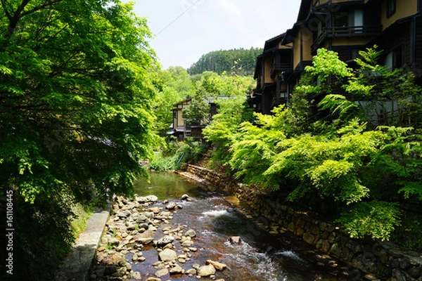 Fototapeta View of fresh stream with stone bank through green trees and local buildings on stone bank in Kurokawa onsen town