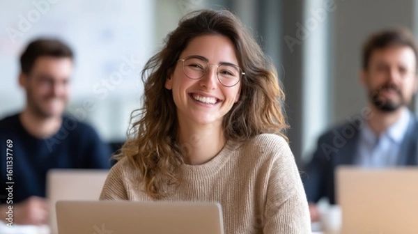 Fototapeta A smiling young woman with glasses sits in front of a laptop, with two blurred men in the background in a bright office setting.