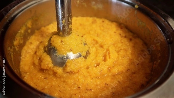 Fototapeta Thick orange vegetable puree is being blended in a metal pot using an immersion hand blender. The hearty texture and warm color suggest a comforting homemade dish