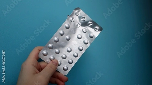 Fototapeta A person holds a silver blister pack of tablets against a blue background. The packaging is partially used, indicating ongoing medication or supplement intake.