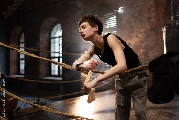 Fototapeta A woman in a boxing ring, wrapping an elastic bandage around her arms, preparing for boxing training. The photo is in a cinematic style, the light gives the mood to a sports motivational story about