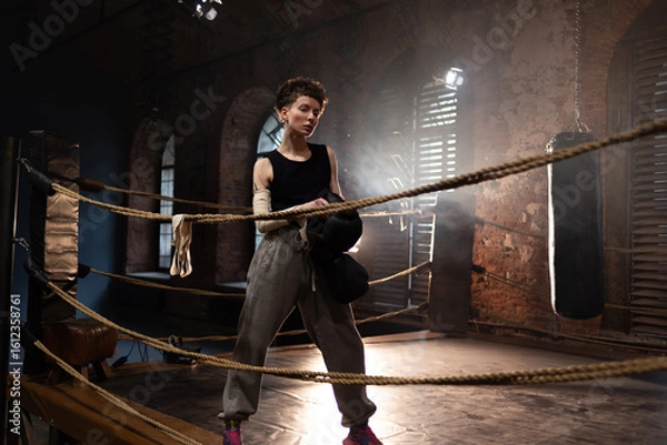 Obraz A woman in a boxing gym, portrait by the ropes, cinematic style, dark tones. Training and overcoming oneself, motivation