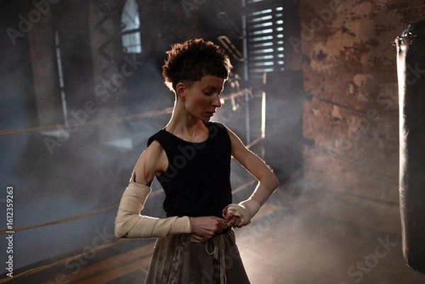 Obraz A woman in a boxing ring, wrapping an elastic bandage around her arms, preparing for boxing training. The photo is in a cinematic style, the light gives the mood to a sports motivational story about
