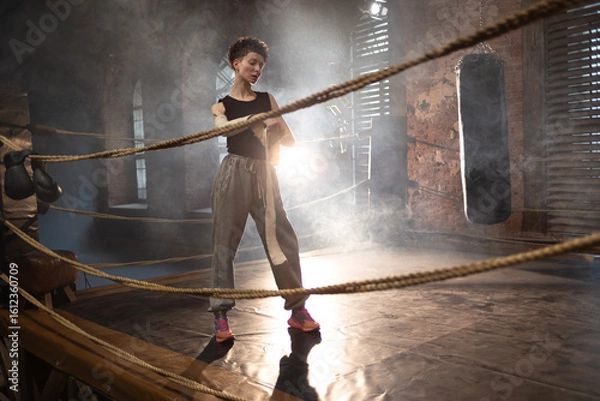Obraz A woman in a boxing ring, wrapping an elastic bandage around her arms, preparing for boxing training. The photo is in a cinematic style, the light gives the mood to a sports motivational story about
