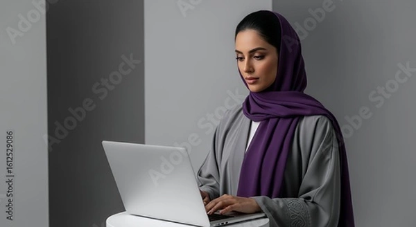 Fototapeta A focused woman in a hijab uses a laptop computer, working diligently from home or in a modern .