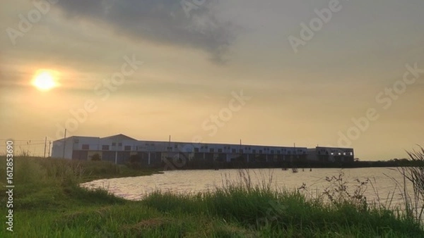 Fototapeta A block of warehouses stands near a quiet pond, bathed in golden hour light. Tall grass sways in the foreground, adding a natural contrast to the industrial setting.