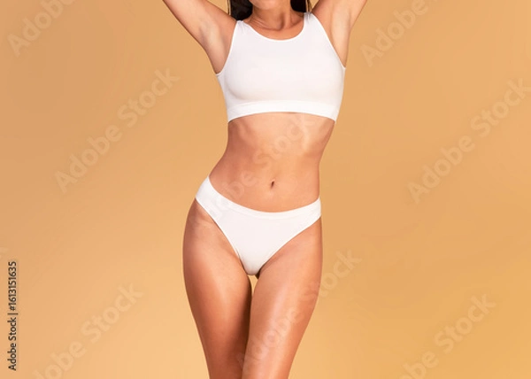 Fototapeta Cropped front view of slender lady with ideal body and flat stomach posing in white underwear on beige studio background, woman with fit figure wearing top bra and panties