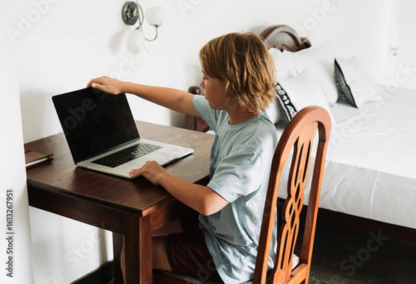 Obraz Teenage boy studying with a laptop in a bright tropical room in Sri Lanka. Digital nomad lifestyle, online learning, and early IT education. Cozy workspace with natural light and wooden furniture
