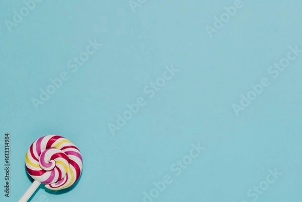 Fototapeta Vibrant pink, yellow, white swirled lollipop on a calming blue background. Perfect for sweetness, childhood, joy, treats, simple pleasures, with copy space.