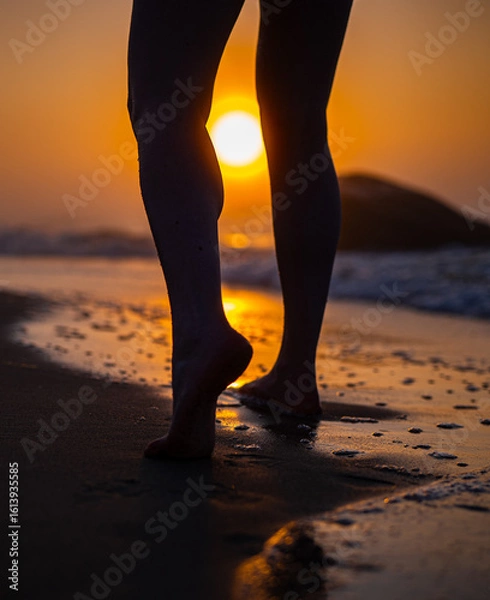 Fototapeta Barefoot person walking on wet sand at beach during sunrise. Close up of legs with ocean waves and glowing sun. Seaside walk and relaxation concept. 