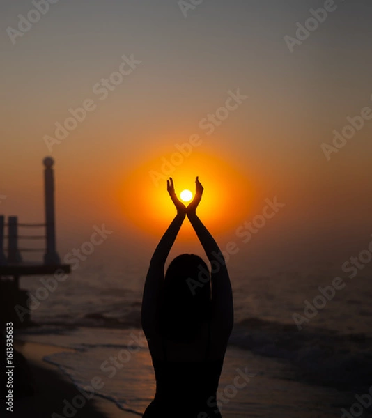 Fototapeta Silhouette of woman standing on beach holding sun with raised hands during sunrise. Ocean shore and pier at morning. Sunsrise and relaxation concept.