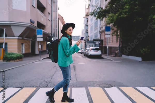 Obraz Confident young woman crossing street while using smartphone, casual green sweater and black hat, exploring city with relaxed attitude and tech-ready mindset.